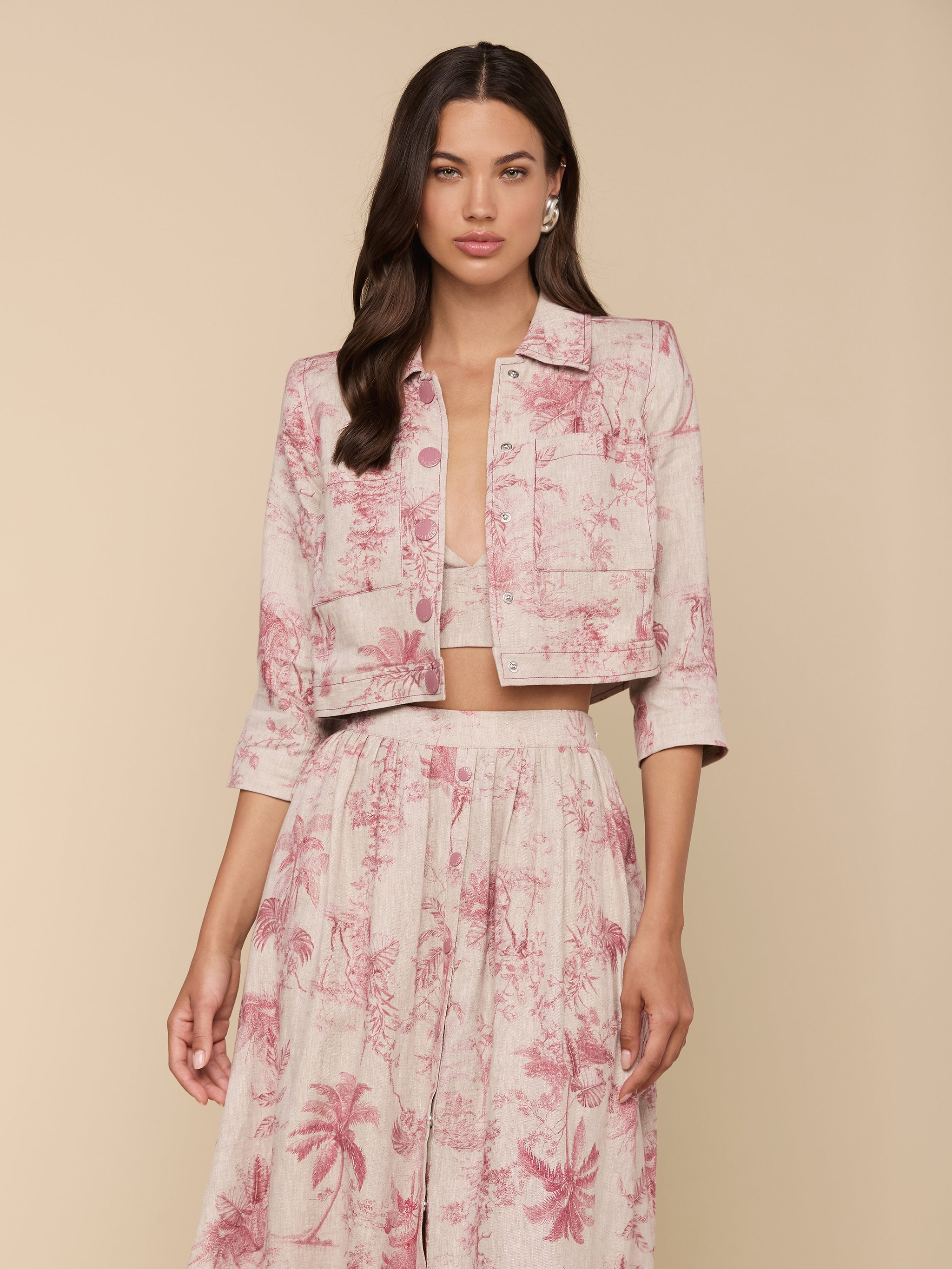Woman wearing the Eda Cropped Linen Jacket in light pink with a red botanical and tropical print, paired with a matching high-waisted skirt, standing against a beige background.
