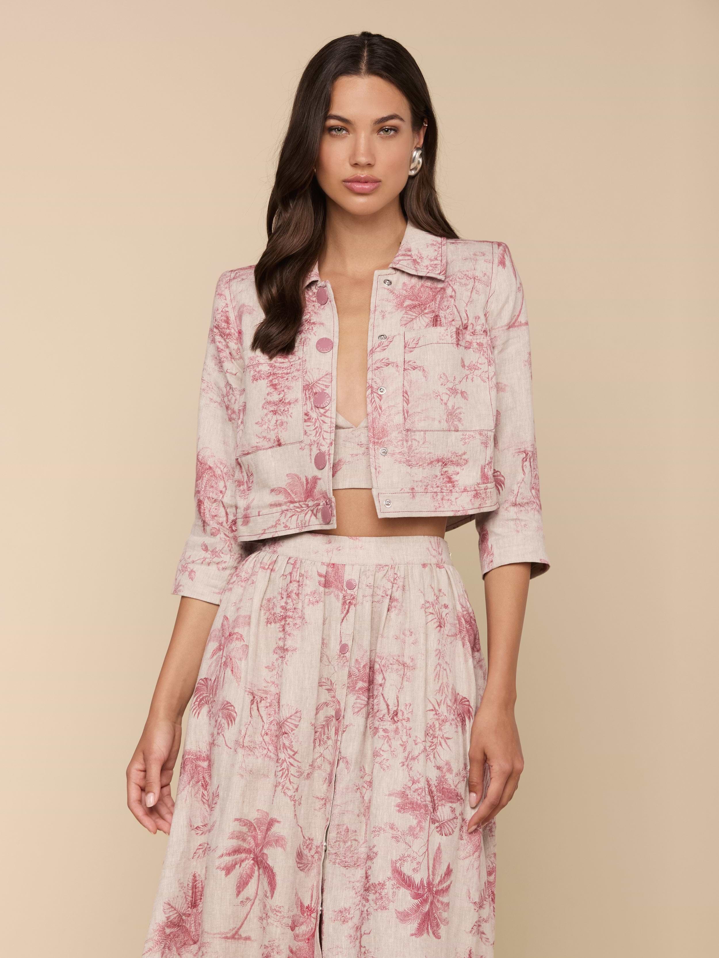 Woman wearing the Eda Cropped Linen Jacket in light pink with a red botanical and tropical print, paired with a matching high-waisted skirt, standing against a beige background.