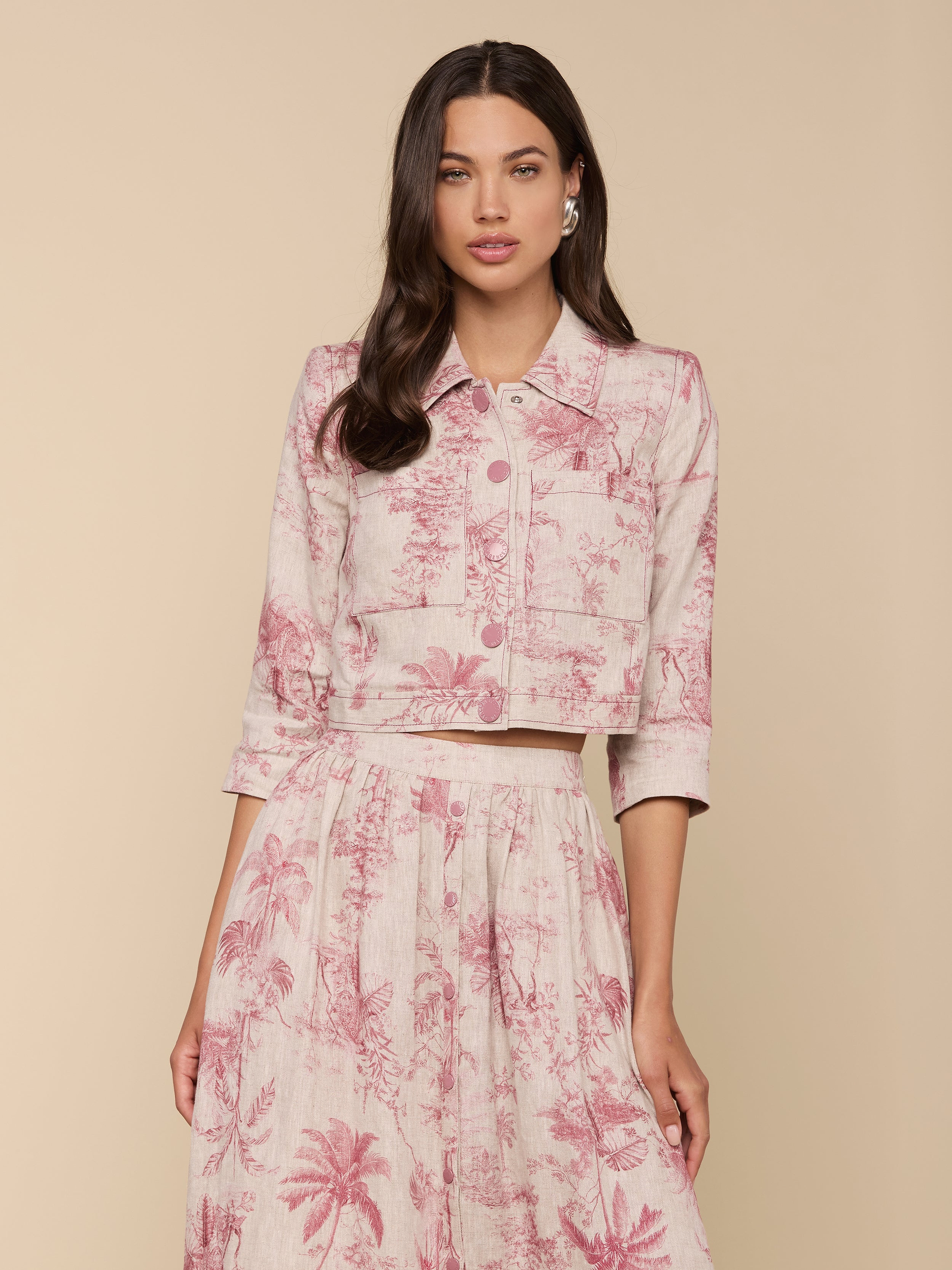 A woman wears the Eda Cropped Linen Jacket, light pink and white patterned, paired with a matching high-waisted skirt, standing against a beige background with her hair down and one hand by her side.