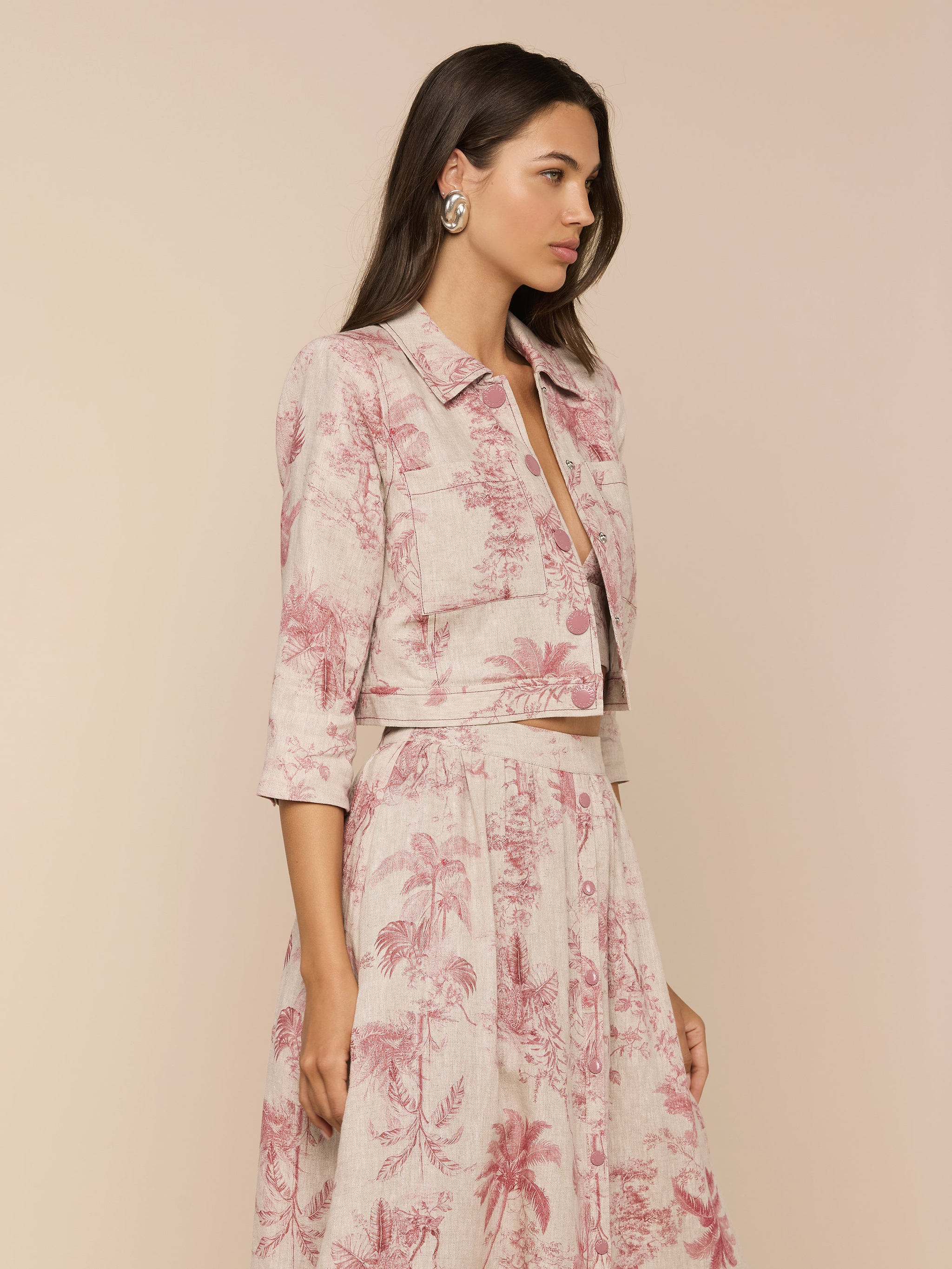 A woman with long brown hair and large earrings models the Eda Cropped Linen Jacket—a light, long-sleeve jacket paired with a matching high-waisted skirt in a pink floral print—standing against a beige background.