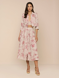 A woman with long dark hair and a neutral expression stands against a beige background, wearing the Eda Cropped Linen Jacket in a light pink and white floral pattern, paired with a matching midi skirt and beige platform heels.