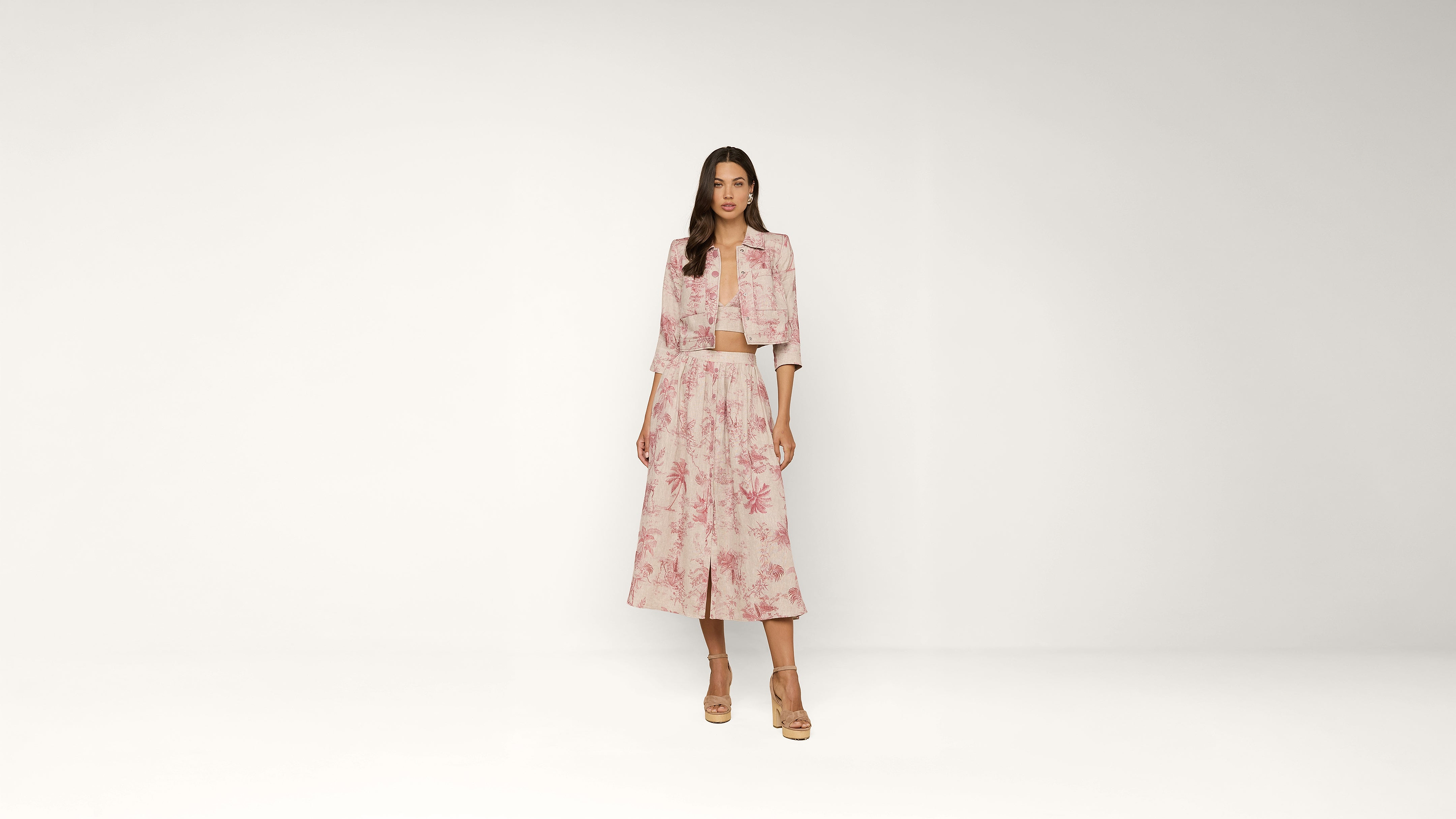 A woman stands against a plain background wearing a light pink floral jacket and matching wide-leg skirt with tan heels.