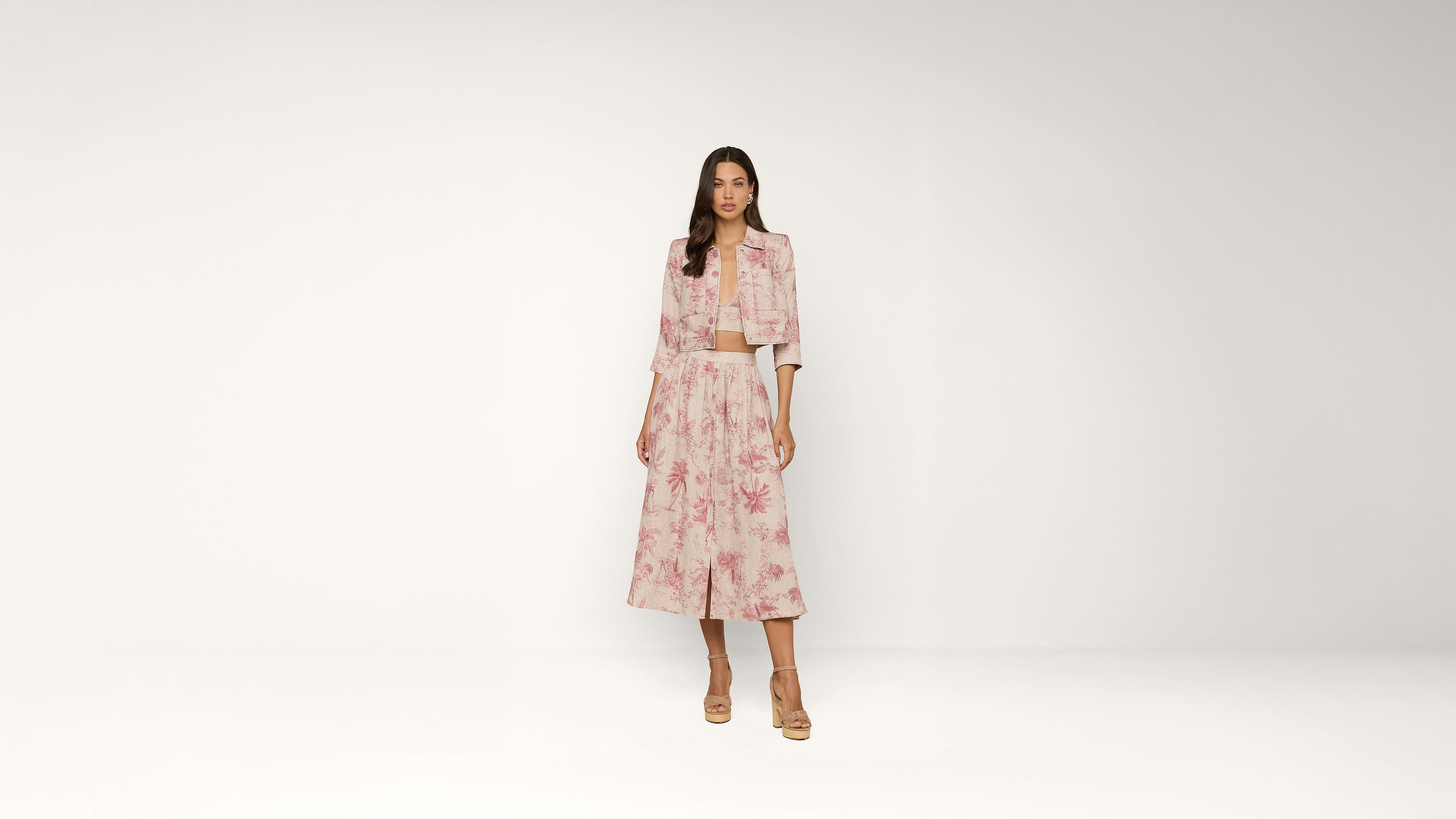 A woman stands against a plain background wearing a light pink floral jacket and matching wide-leg skirt with tan heels.