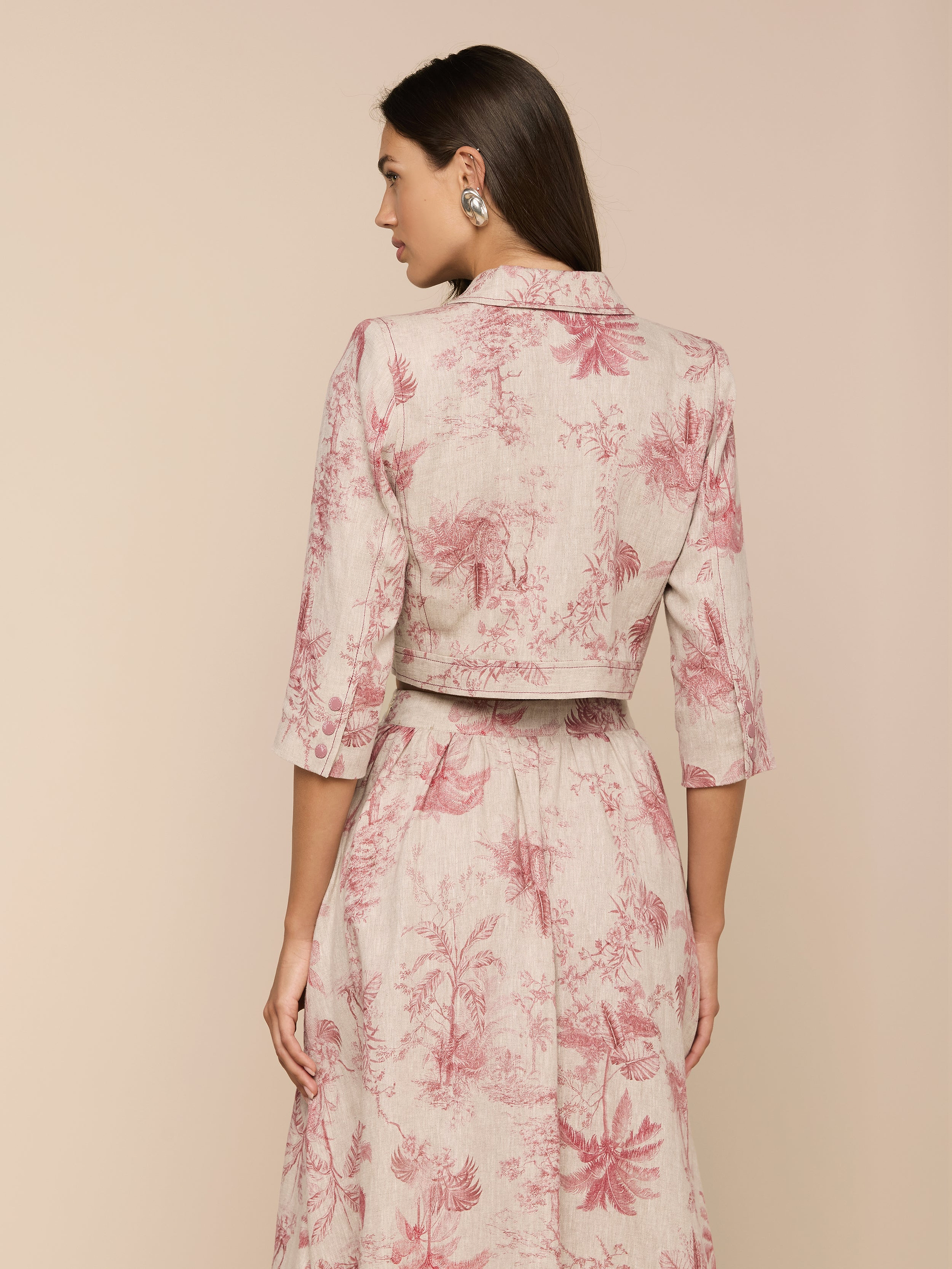 A woman with long dark hair and large earrings stands facing away, wearing the Eda Cropped Linen Jacket—a light-colored, collared top with three-quarter sleeves and pink botanical prints—paired with a long, flowing skirt.