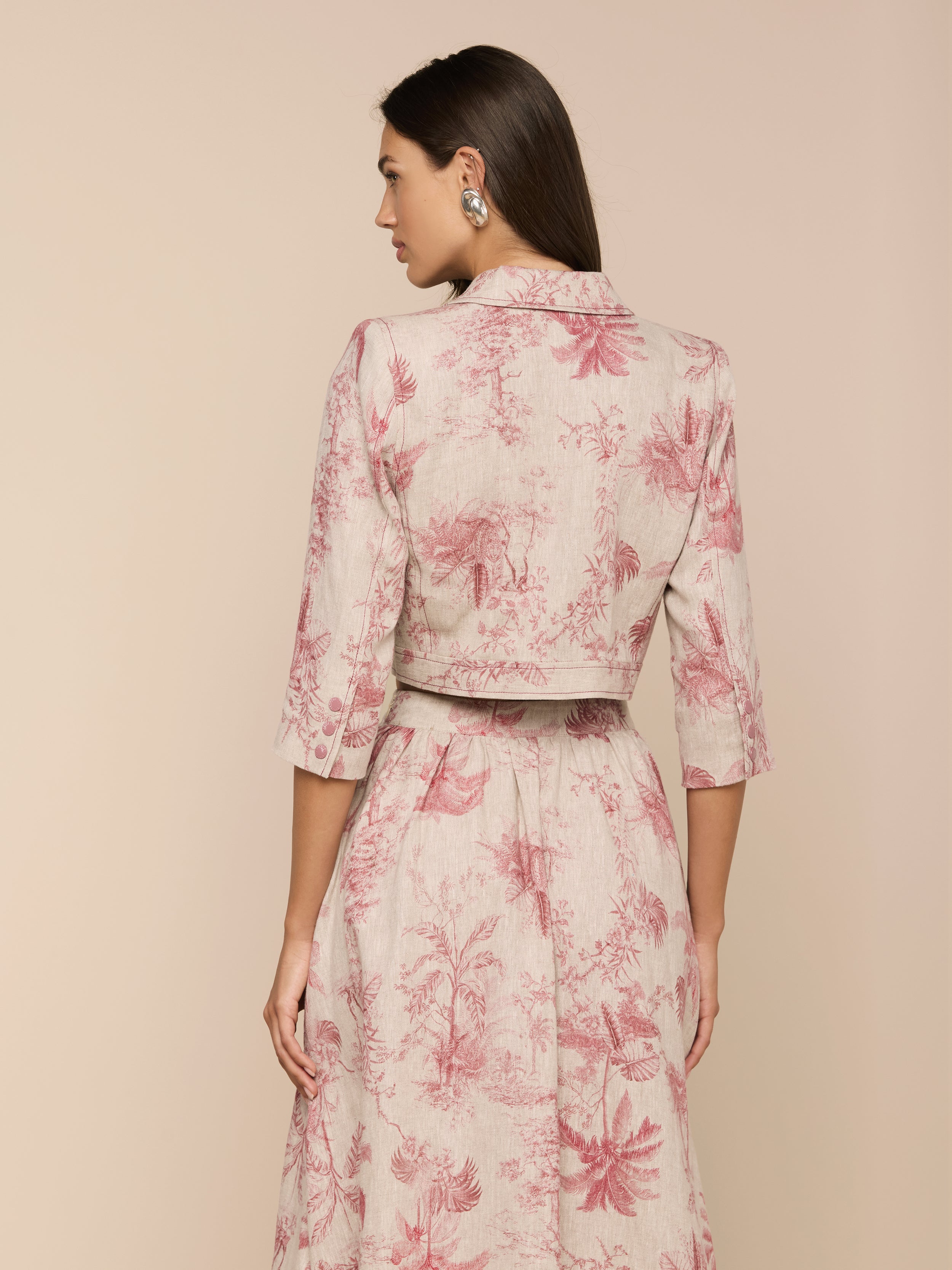 A woman with long dark hair and large earrings stands facing away, wearing the Eda Cropped Linen Jacket—a light-colored, collared top with three-quarter sleeves and pink botanical prints—paired with a long, flowing skirt.