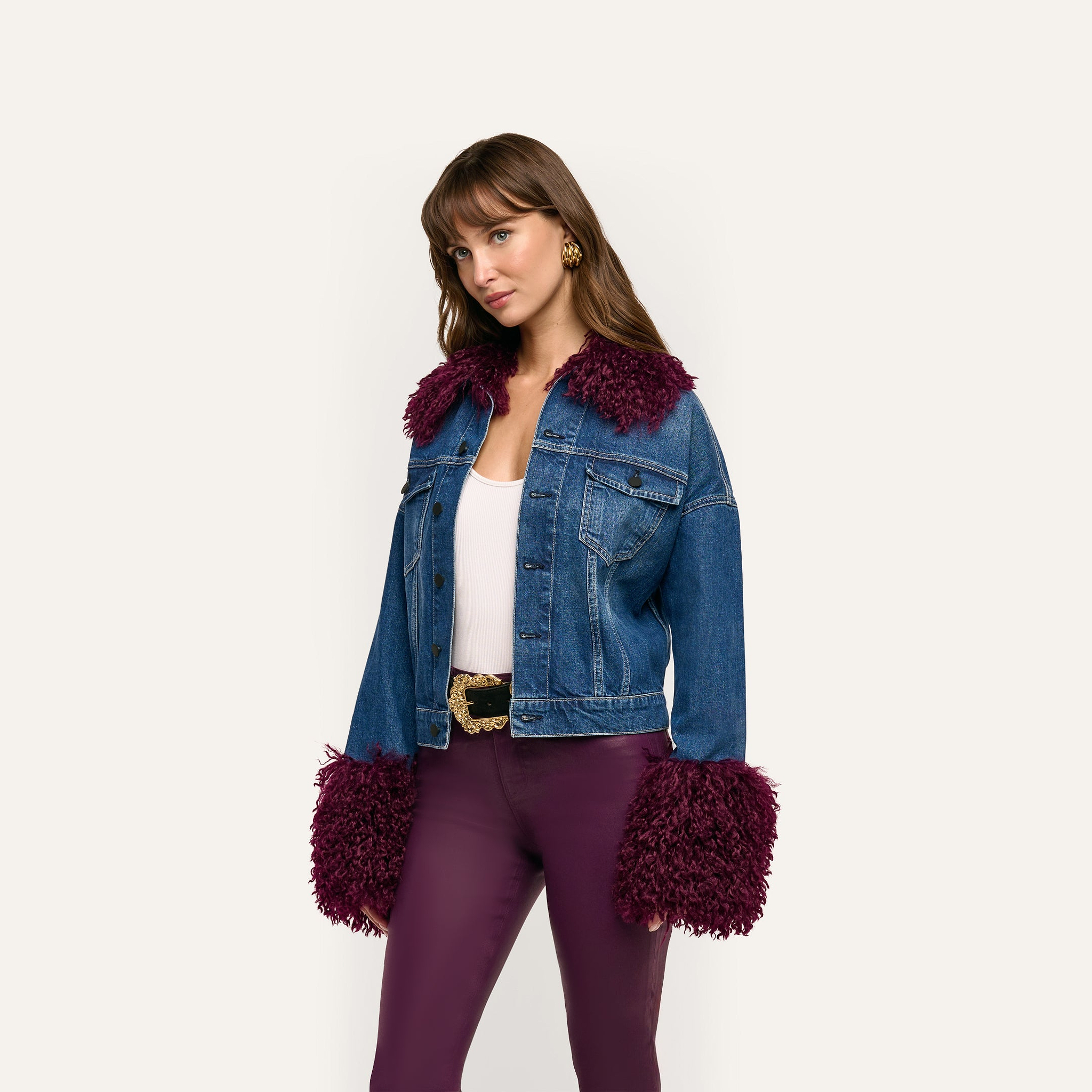 A person wears a Bronson Jean Jacket, a cropped blue denim style with dark purple faux-fur trim on the collar and cuffs, over a white tank top and dark purple trousers.