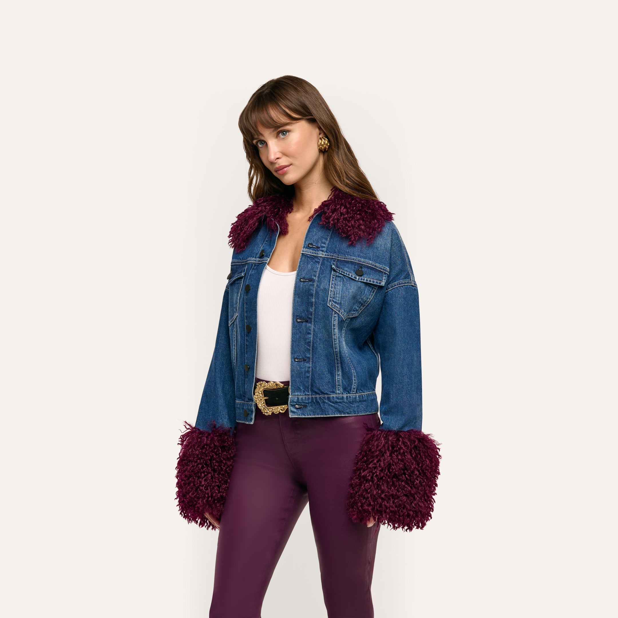 A person wears a Bronson Jean Jacket, a cropped blue denim style with dark purple faux-fur trim on the collar and cuffs, over a white tank top and dark purple trousers.