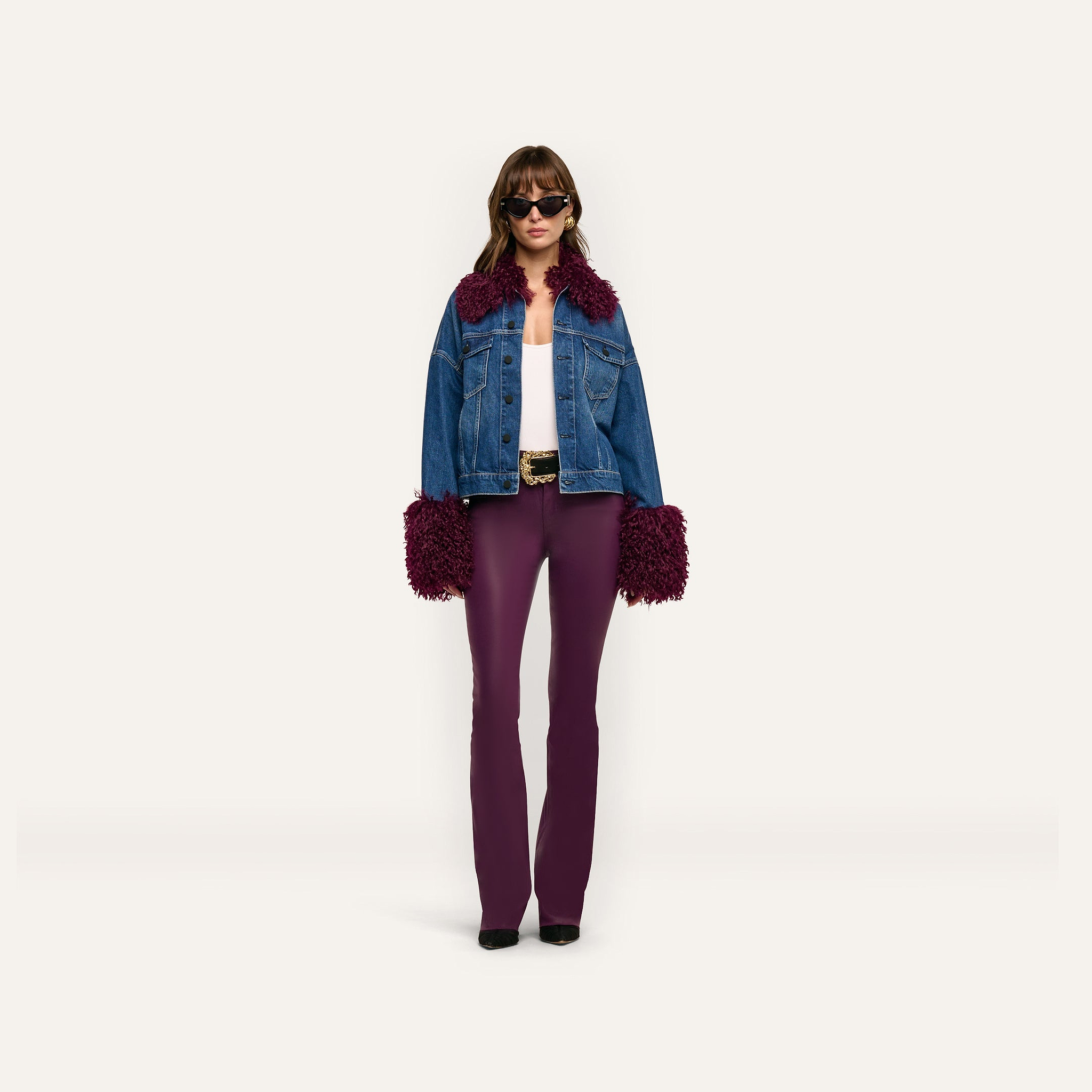 A woman wears a Bronson Jean Jacket featuring a cropped blue denim design with dark purple faux-fur trim on the collar and cuffs, paired with a white top and fitted purple trousers.