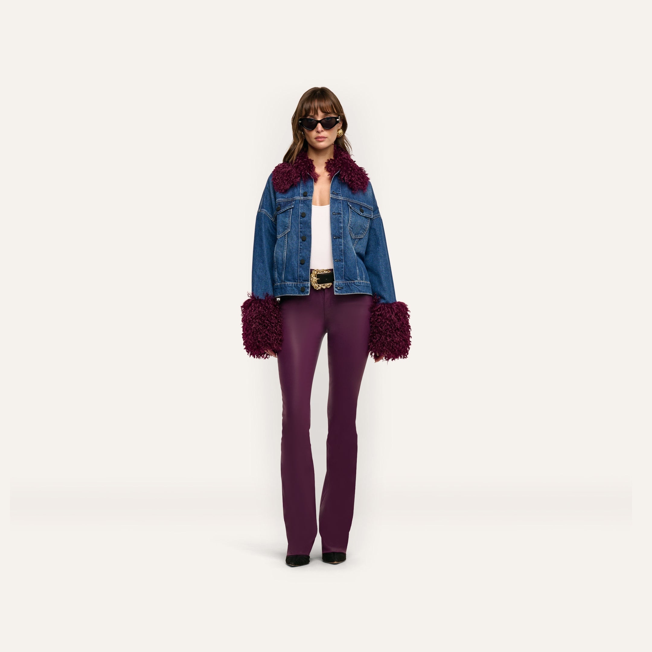 A woman wears a Bronson Jean Jacket featuring a cropped blue denim design with dark purple faux-fur trim on the collar and cuffs, paired with a white top and fitted purple trousers.