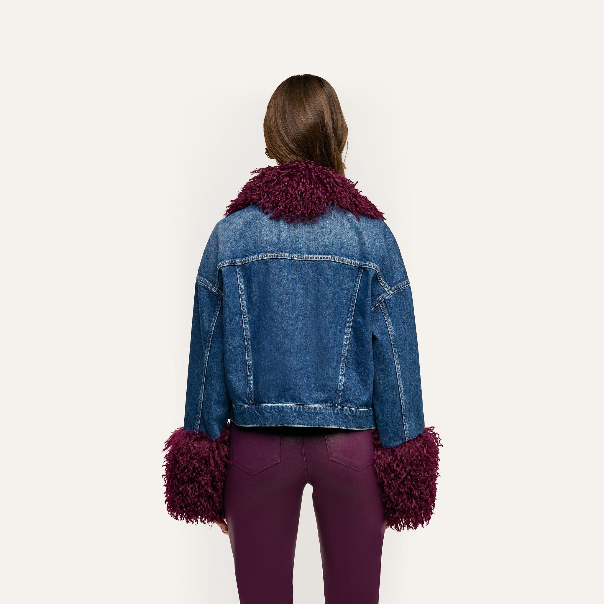 Back view of a Bronson Jean Jacket, a cropped blue denim jacket with dark purple, shaggy faux fur trim on the collar and cuffs, worn over matching purple pants.