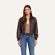 Woman standing against a beige background wearing the Clotilde Leather Jacket, a dark brown cropped bomber crafted from fine-grain leather, paired with a taupe top, blue jeans, hoop earrings, and a brown belt with keys attached. She has long brown hair and a neutral expression, effortlessly showcasing luxury layering.