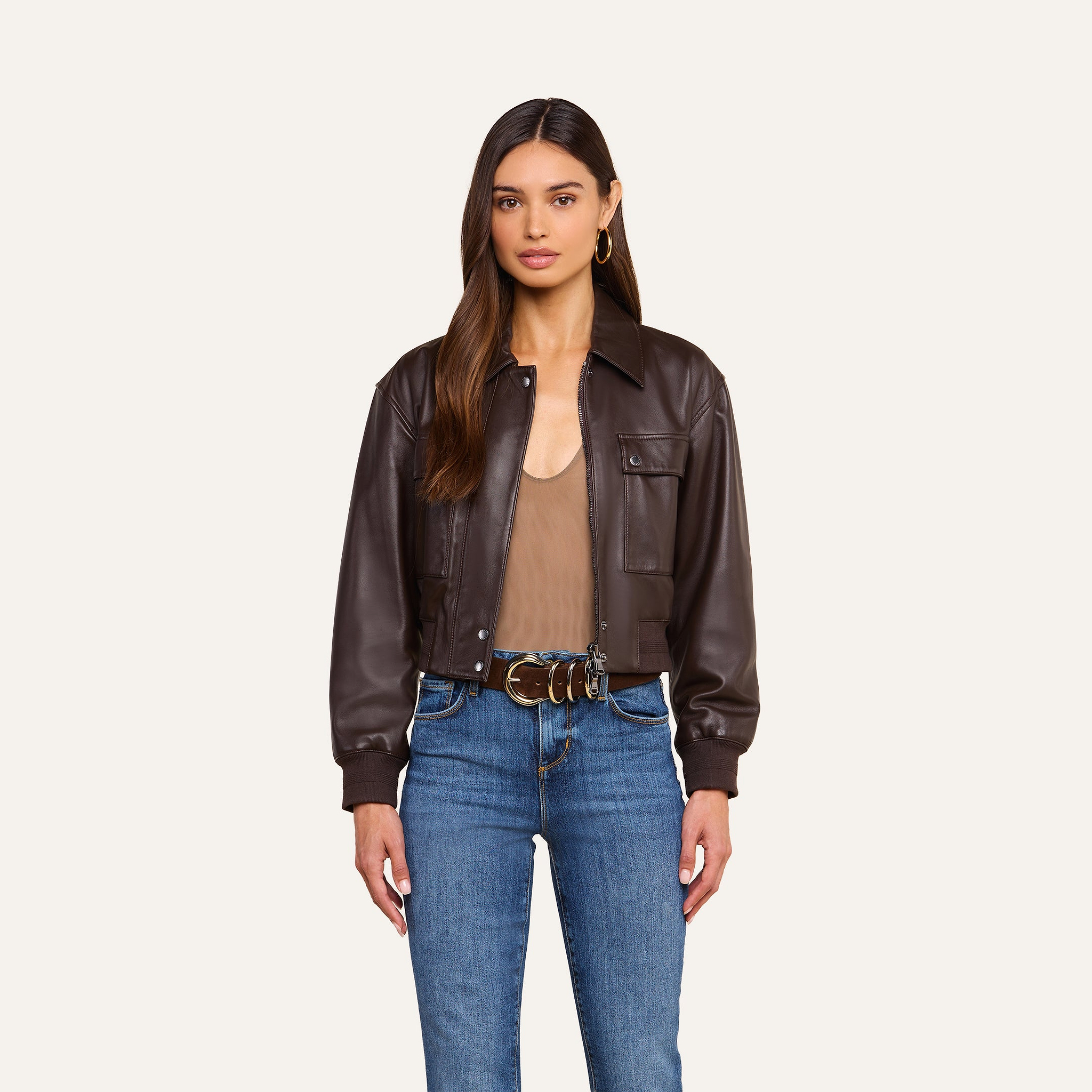 Woman standing against a beige background wearing the Clotilde Leather Jacket, a dark brown cropped bomber crafted from fine-grain leather, paired with a taupe top, blue jeans, hoop earrings, and a brown belt with keys attached. She has long brown hair and a neutral expression, effortlessly showcasing luxury layering.