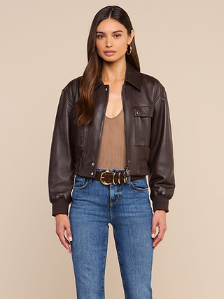 Woman standing against a beige background wearing the Clotilde Leather Jacket, a dark brown cropped bomber crafted from fine-grain leather, paired with a taupe top, blue jeans, hoop earrings, and a brown belt with keys attached. She has long brown hair and a neutral expression, effortlessly showcasing luxury layering.