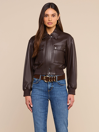 A woman wearing the Clotilde Leather Jacket in dark brown fine-grain leather, paired with blue jeans and a brown belt featuring a gold buckle, stands against a plain beige background—showcasing luxury layering at its finest.