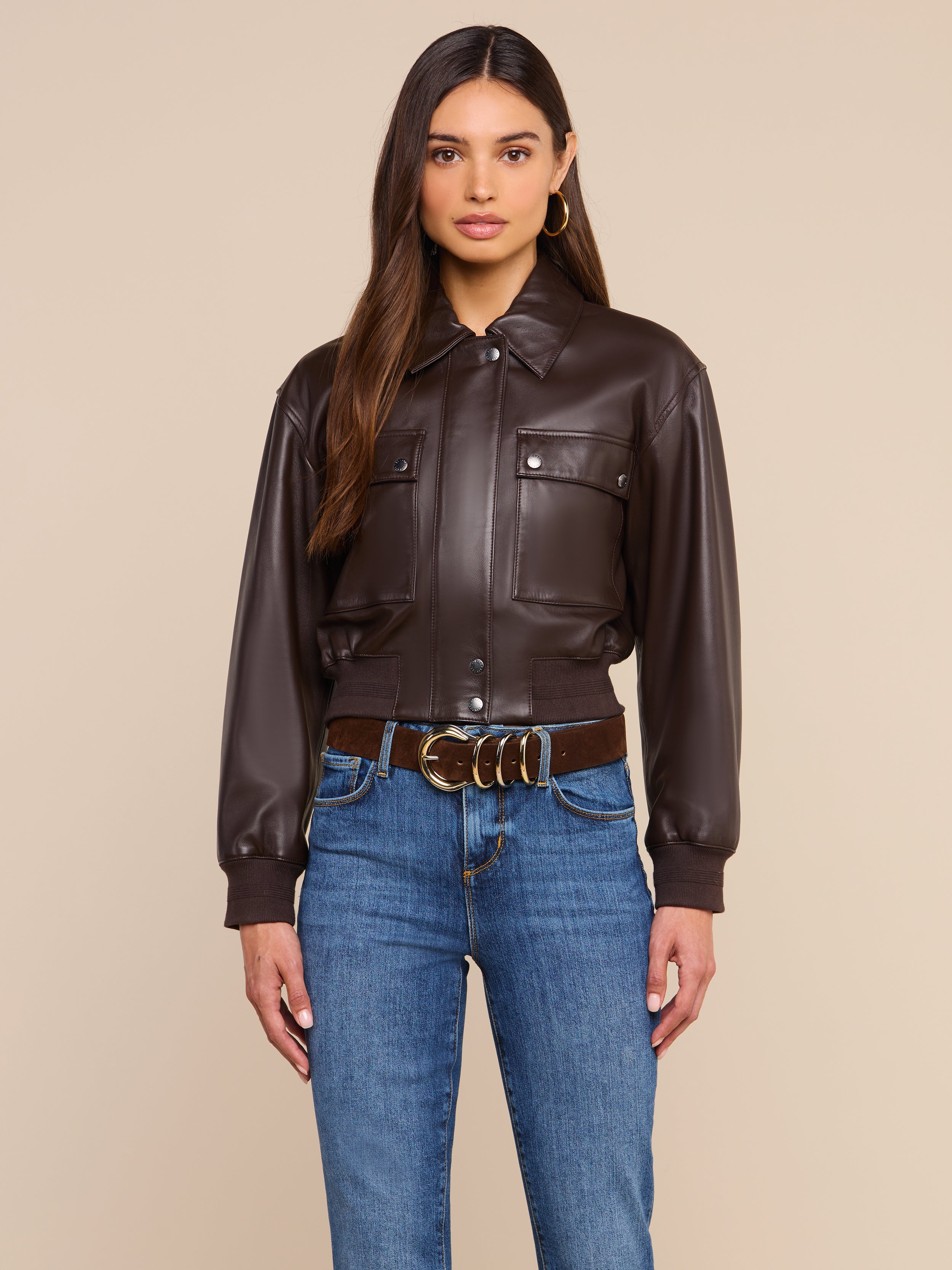 A woman wearing the Clotilde Leather Jacket in dark brown fine-grain leather, paired with blue jeans and a brown belt featuring a gold buckle, stands against a plain beige background—showcasing luxury layering at its finest.
