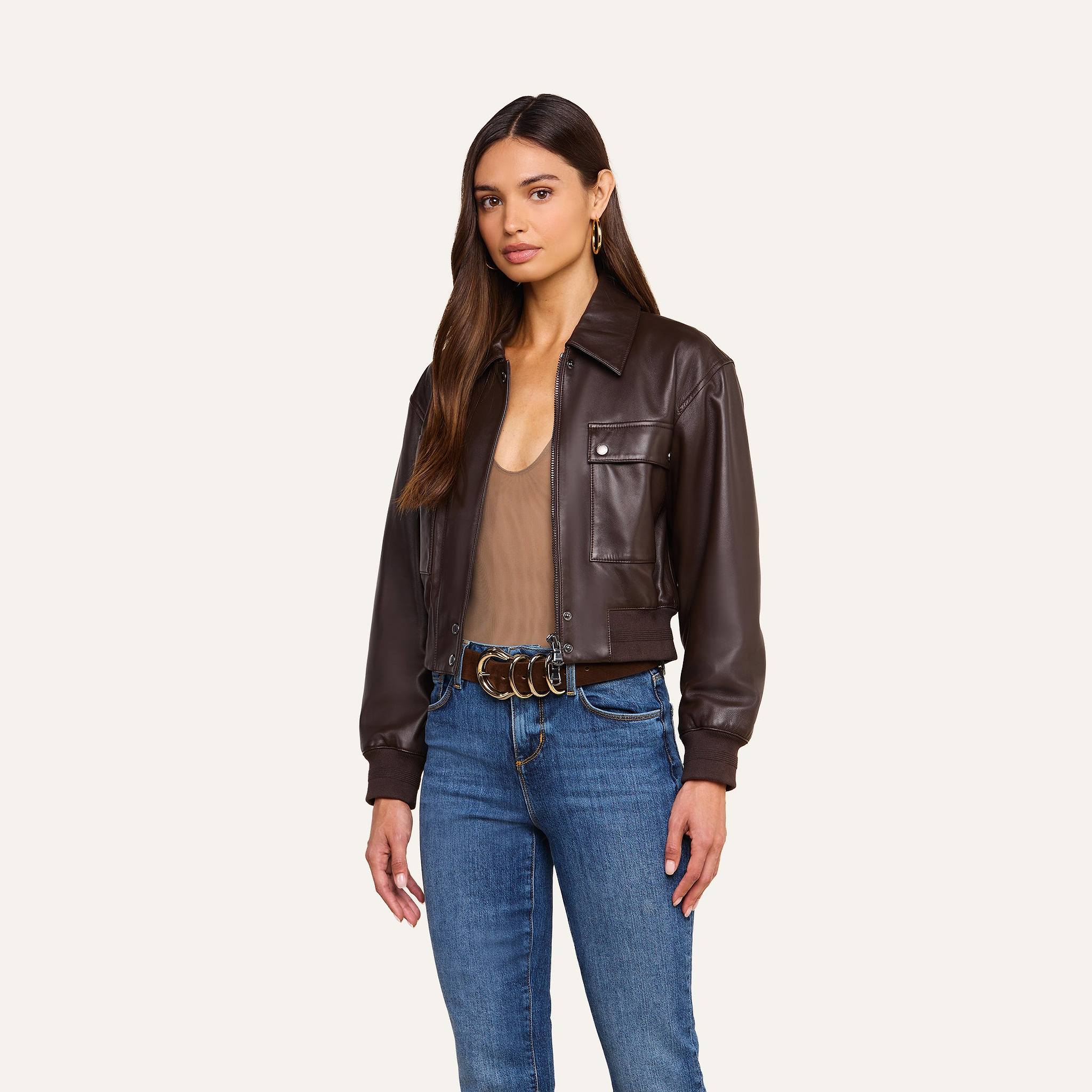 A woman stands against a plain beige background, effortlessly embracing the art of luxury layering in the Clotilde Leather Jacket—a fine-grain brown leather piece—paired with a tan top, blue jeans, and a belt with gold rings.