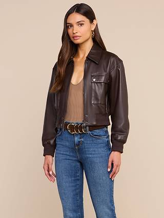 A woman stands against a plain beige background, effortlessly embracing the art of luxury layering in the Clotilde Leather Jacket—a fine-grain brown leather piece—paired with a tan top, blue jeans, and a belt with gold rings.