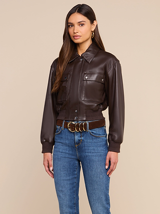 Clotilde Leather Jacket: Woman with long brown hair styled in the Clotilde Leather Jacket, paired with blue jeans, a brown belt featuring a gold buckle, and gold hoop earrings, standing against a plain beige background—a perfect ensemble for effortless luxury layering.