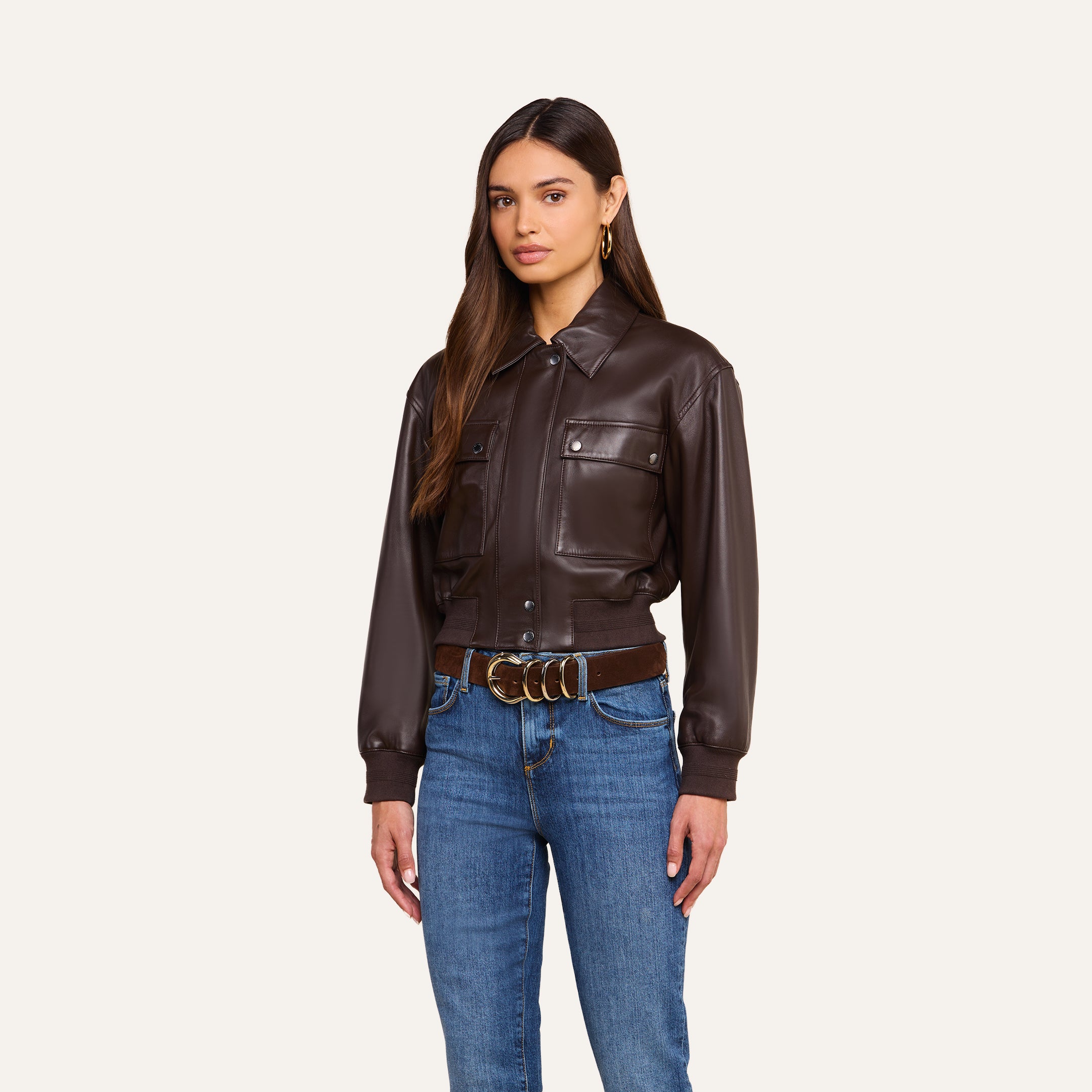 Clotilde Leather Jacket: Woman with long brown hair styled in the Clotilde Leather Jacket, paired with blue jeans, a brown belt featuring a gold buckle, and gold hoop earrings, standing against a plain beige background—a perfect ensemble for effortless luxury layering.