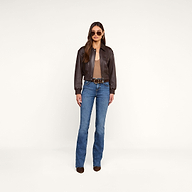 A woman wearing sunglasses, the Clotilde Leather Jacket—a fine-grain leather cropped bomber—over a sheer top, blue high-waisted jeans with a belt, and brown boots stands against a plain beige background.