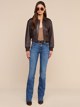 A woman wearing sunglasses, the Clotilde Leather Jacket—a fine-grain leather cropped bomber—over a sheer top, blue high-waisted jeans with a belt, and brown boots stands against a plain beige background.
