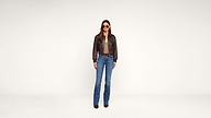 A woman stands facing forward, wearing sunglasses, a brown leather jacket, a brown shirt, blue jeans, and brown shoes, against a plain white background.