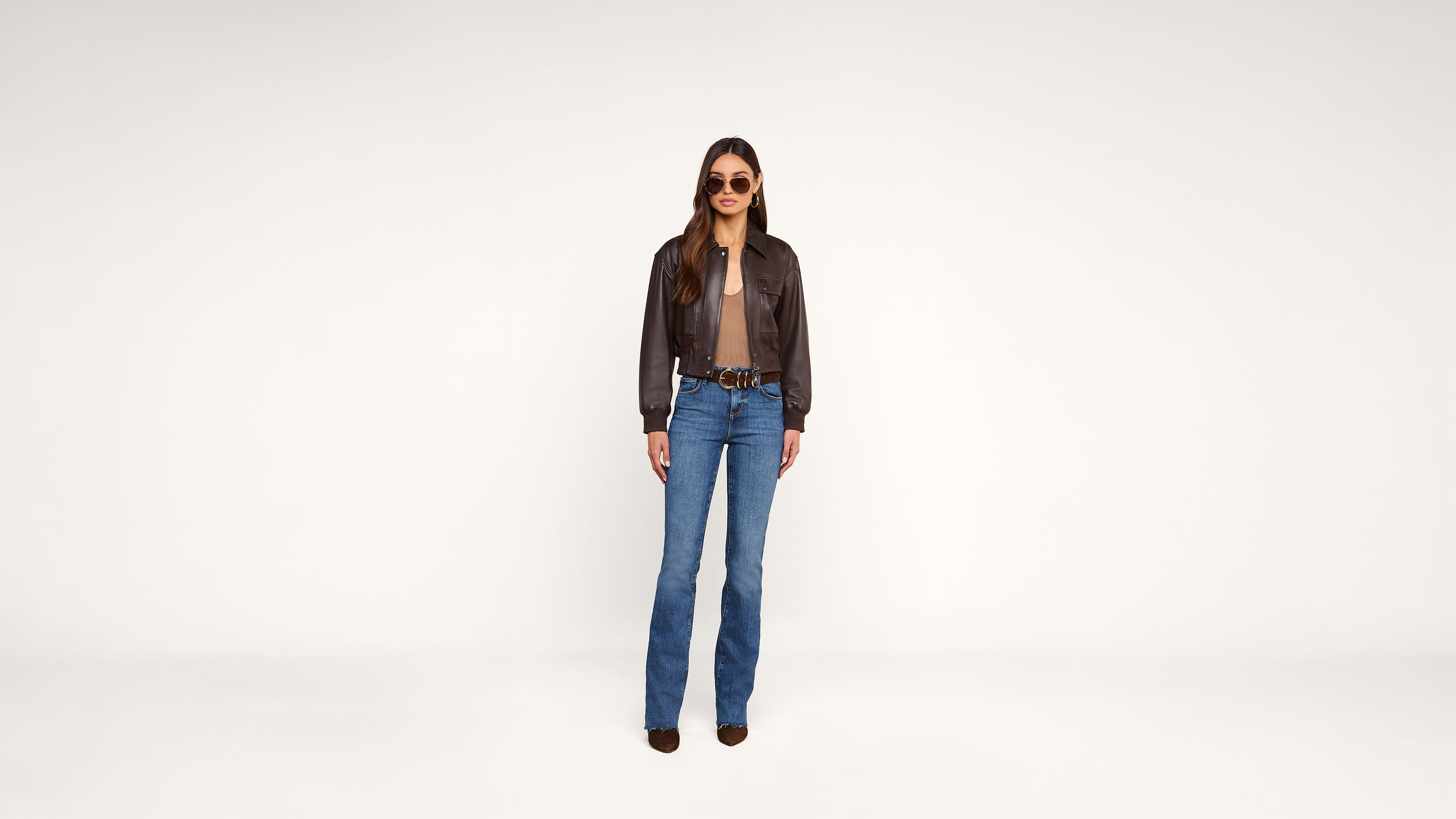 A woman stands facing forward, wearing sunglasses, a brown leather jacket, a brown shirt, blue jeans, and brown shoes, against a plain white background.