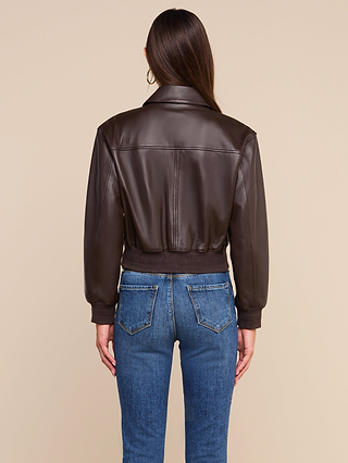 A woman stands facing away, wearing the Clotilde Leather Jacket—crafted from fine-grain leather—and blue jeans against a plain beige background, offering a perfect example of luxury layering.