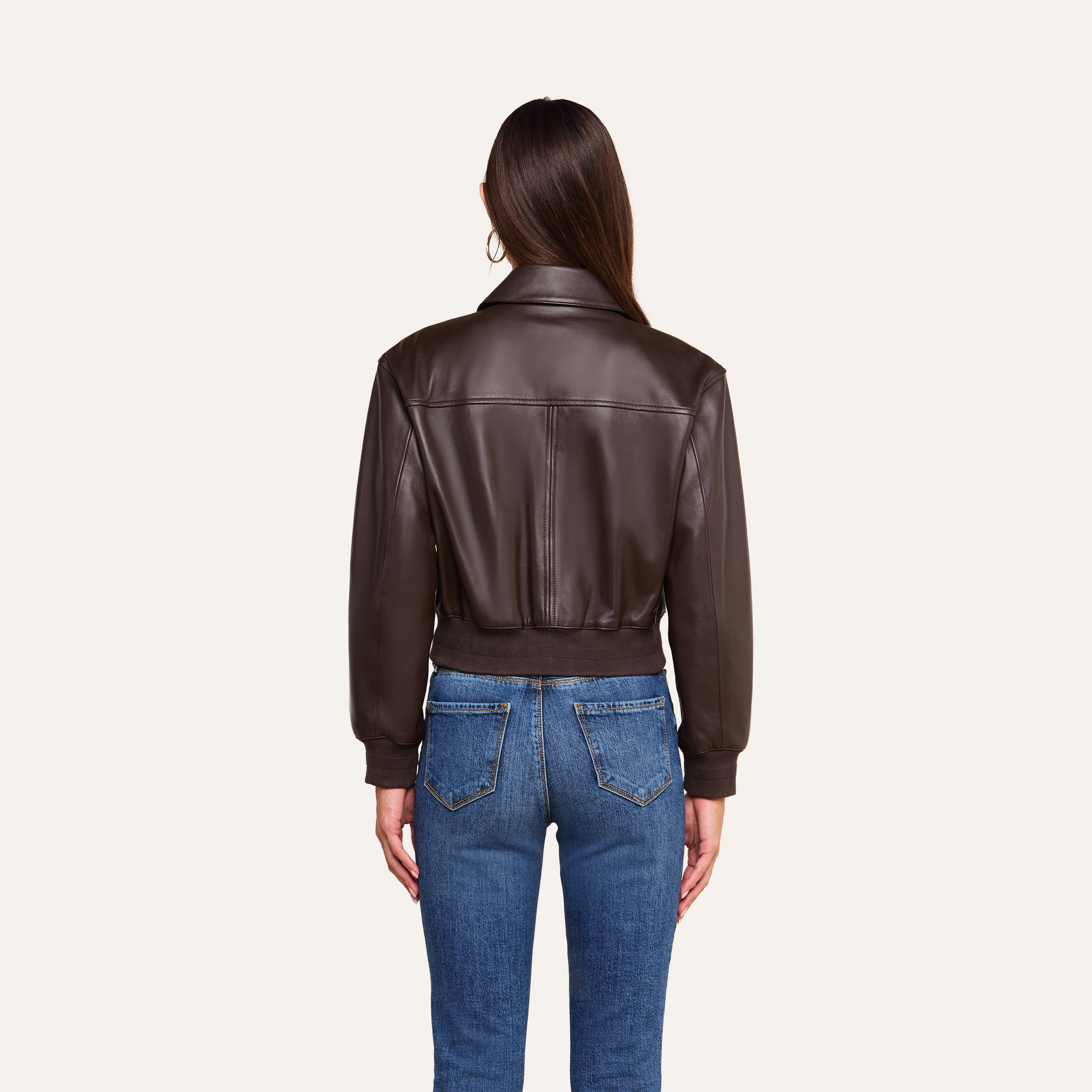 A woman stands facing away, wearing the Clotilde Leather Jacket—crafted from fine-grain leather—and blue jeans against a plain beige background, offering a perfect example of luxury layering.