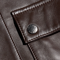 Close-up of dark brown, fine-grain leather fabric with visible grain, showing a flap secured by a silver snap button. The stitching and texture of the Clotilde Leather Jacket’s luxury layering material are clearly detailed, reminiscent of what you’d find on a cropped bomber jacket.
