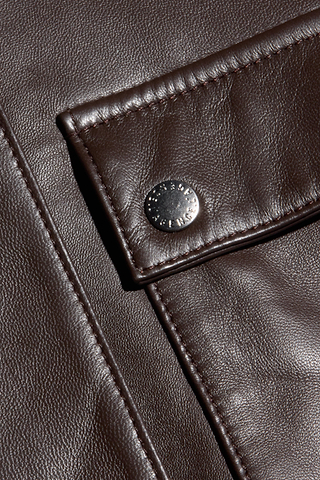 Close-up of dark brown, fine-grain leather fabric with visible grain, showing a flap secured by a silver snap button. The stitching and texture of the Clotilde Leather Jacket’s luxury layering material are clearly detailed, reminiscent of what you’d find on a cropped bomber jacket.