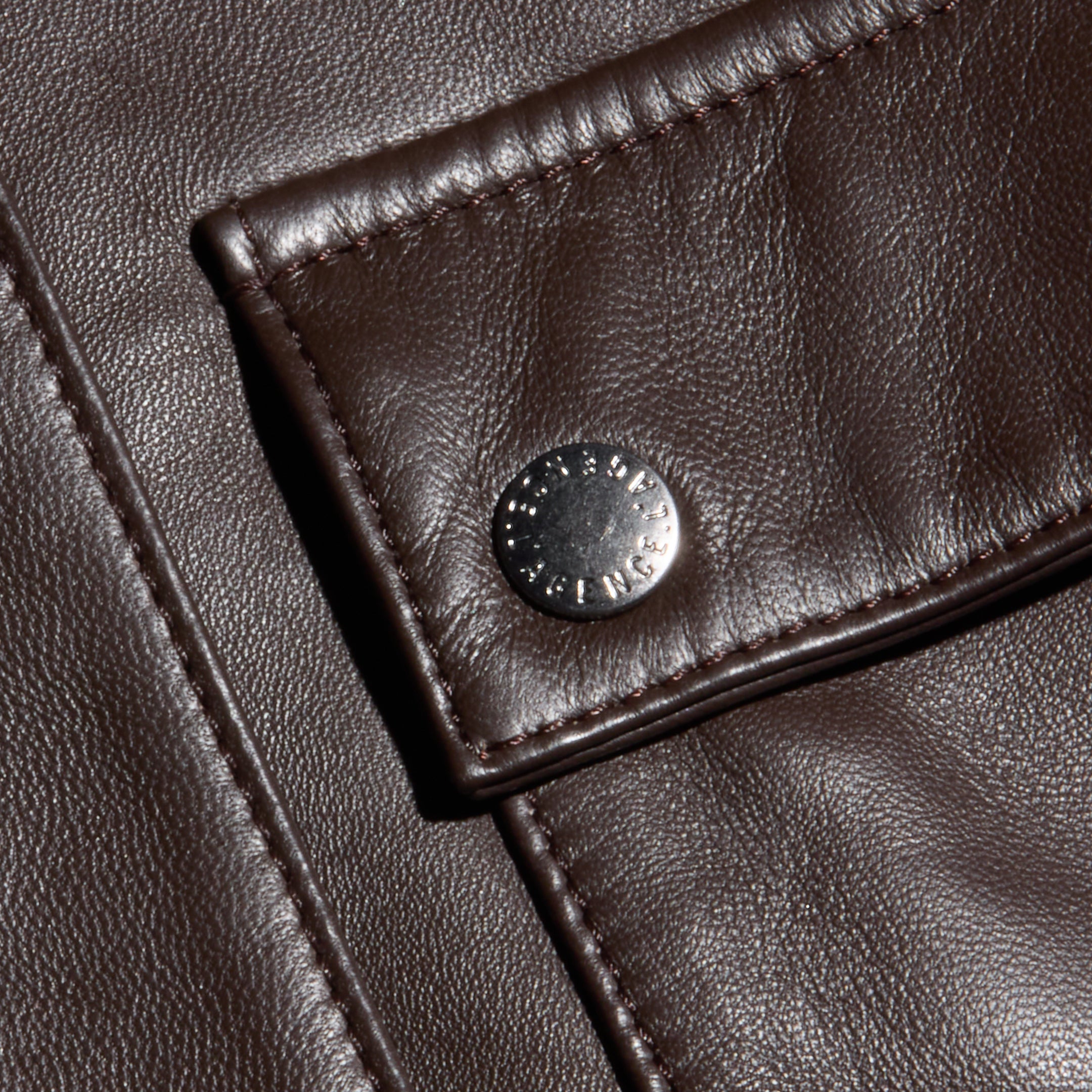 Close-up of dark brown, fine-grain leather fabric with visible grain, showing a flap secured by a silver snap button. The stitching and texture of the Clotilde Leather Jacket’s luxury layering material are clearly detailed, reminiscent of what you’d find on a cropped bomber jacket.