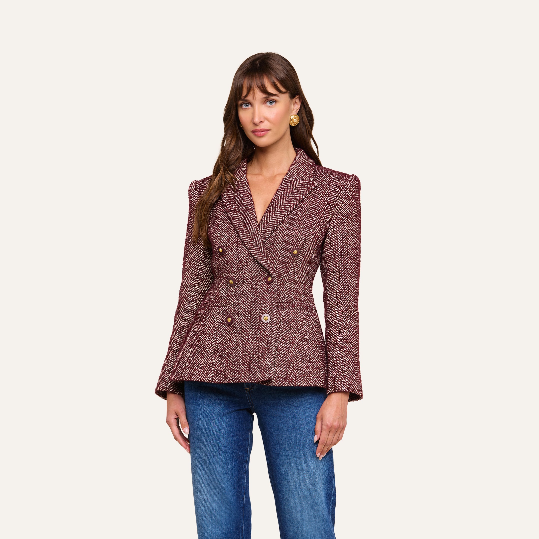 A woman wears the Karima Tweed Blazer, a red herringbone double-breasted jacket with gold buttons, paired with blue jeans and standing against a plain light background.