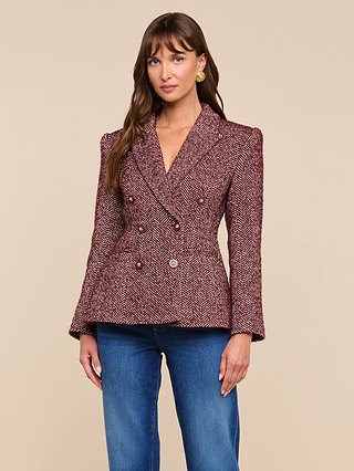 A woman stands against a beige background, wearing the Karima Tweed Blazer—a maroon herringbone tweed blazer with gold buttons—paired with blue jeans. She has long brown hair, gold earrings, and a slight smile.