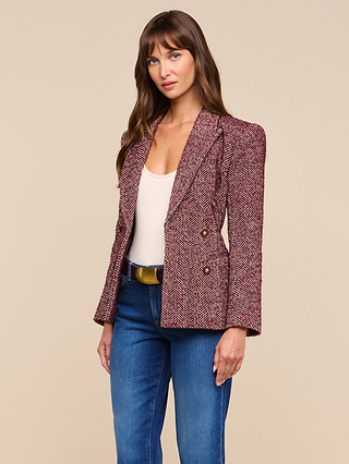 A woman with long brown hair wears the Karima Tweed Blazer in maroon herringbone, paired with a white top, blue jeans, and a brown belt as she stands against a plain beige background. The Karima Tweed Blazer’s classic silhouette adds sophistication to her effortlessly chic look.
