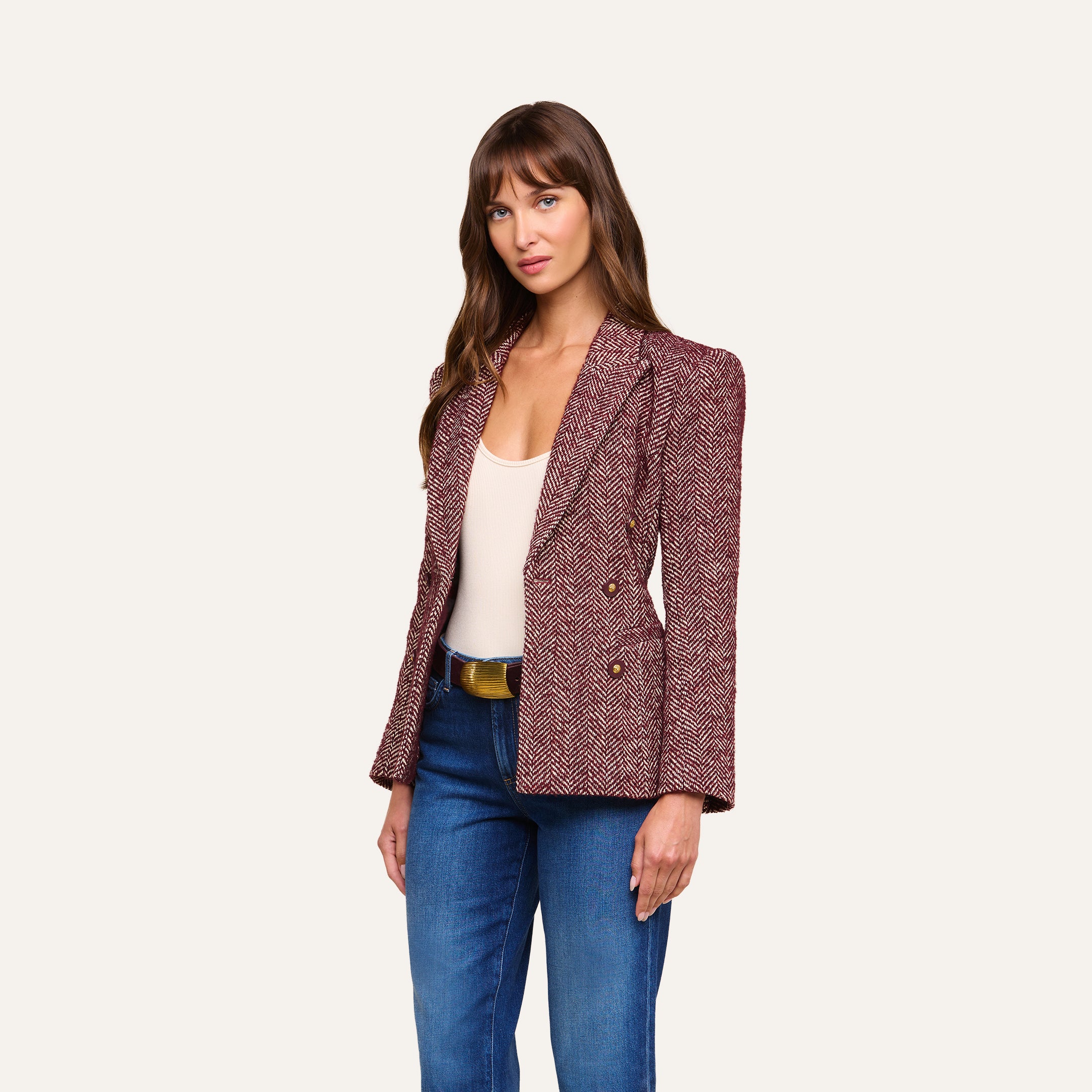 A woman stands against a plain background, looking forward with relaxed posture and straight brown hair, wearing the Karima Tweed Blazer in red over a white tank top and blue jeans.