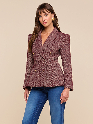 A woman stands against a beige background, looking at the camera with a slight smile, wearing the Karima Tweed Blazer in maroon herringbone with gold buttons, paired with blue jeans.