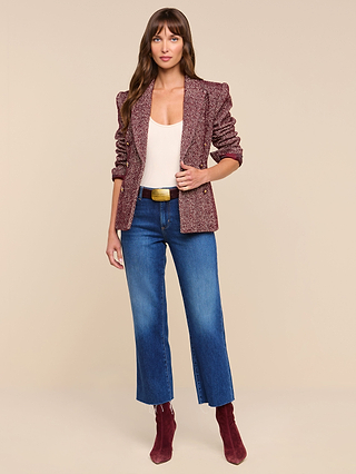 A woman stands against a beige background, wearing the Karima Tweed Blazer in maroon over a cream top, paired with blue cropped jeans, maroon ankle boots, and a belt. She has long brown hair and her hands are relaxed by her side.