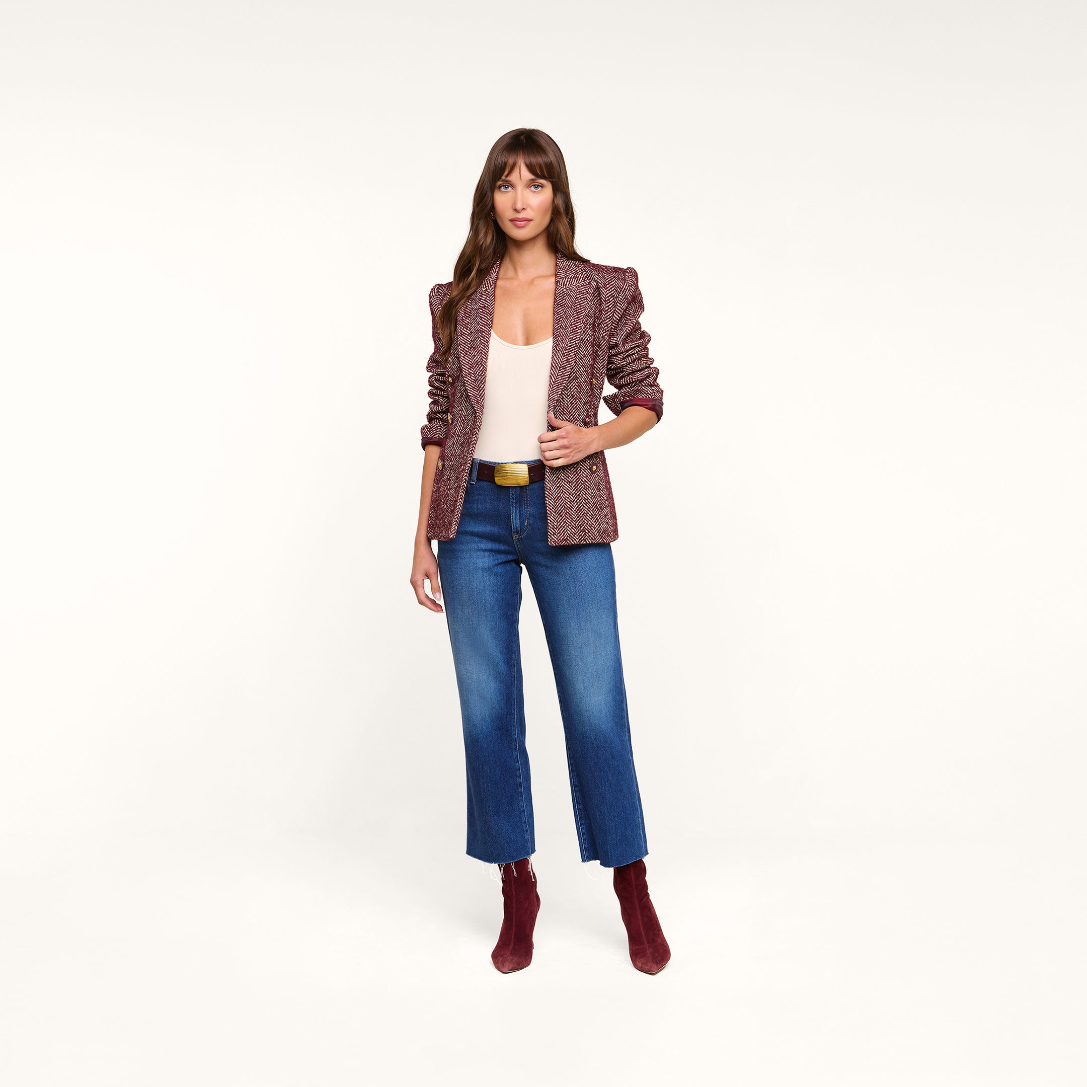 A woman with long brown hair and bangs stands against a white background, wearing the Karima Tweed Blazer in maroon over a white top, paired with blue cropped jeans, maroon ankle boots, and a black belt, looking toward the camera.