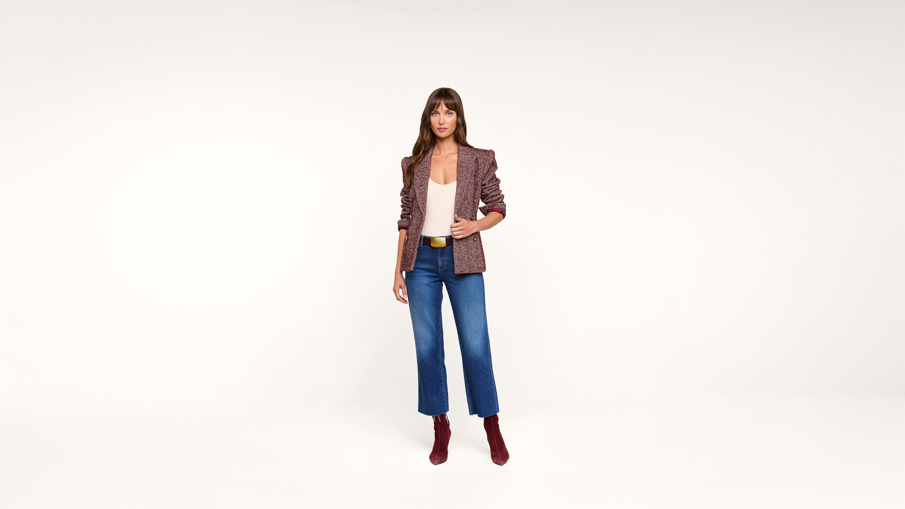 A woman stands in front of a white background, wearing a maroon blazer, white top, blue jeans, and maroon ankle boots.