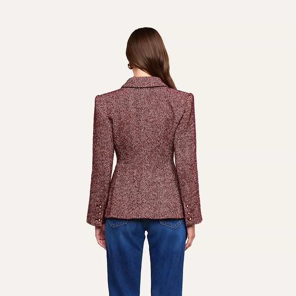 A woman stands facing away, wearing the Karima Tweed Blazer—a fitted maroon herringbone jacket with gold buttons on the sleeves—paired with blue jeans. Her straight brown hair falls down her back, and the background is plain and light-colored.