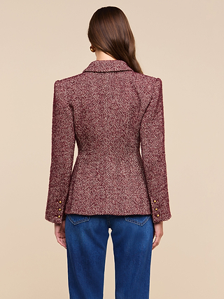 A woman with long brown hair stands with her back to the camera against a beige background, wearing the Karima Tweed Blazer in maroon herringbone and blue jeans, highlighting the classic style of this blazer.