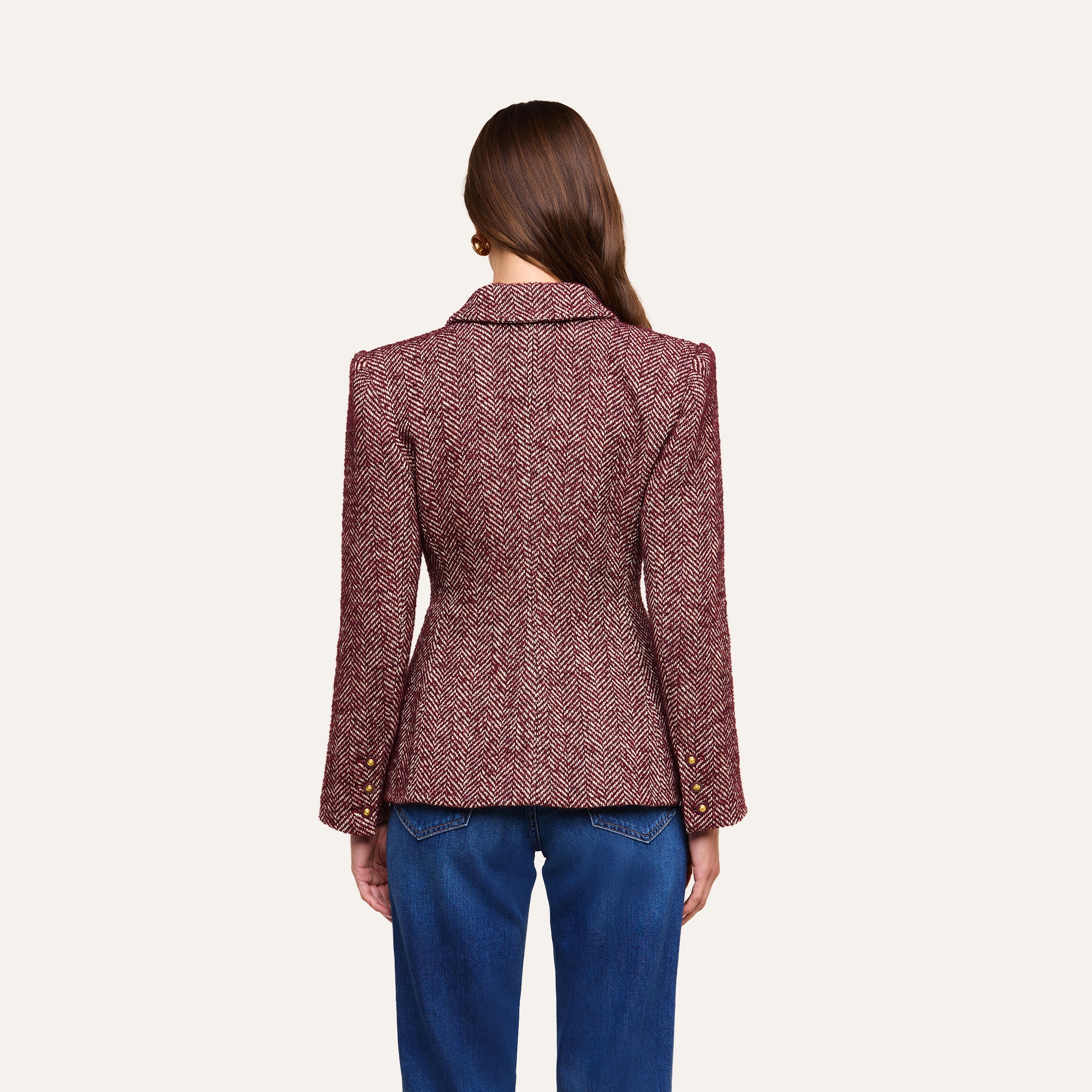 A woman stands facing away, wearing the Karima Tweed Blazer—a fitted maroon herringbone jacket with gold buttons on the sleeves—paired with blue jeans. Her straight brown hair falls down her back, and the background is plain and light-colored.