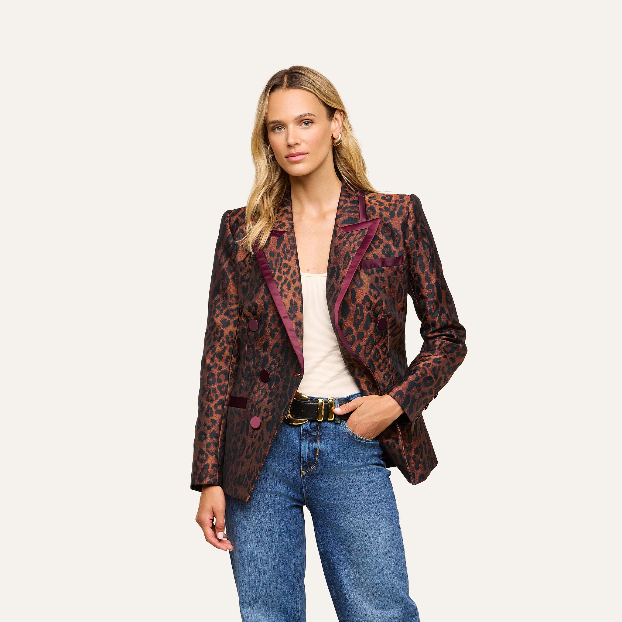 A woman wearing the Kyomi Double-Breasted Blazer with a leopard jacquard pattern and burgundy accents, paired with a white top, blue jeans, and a black belt, stands against a plain light background.