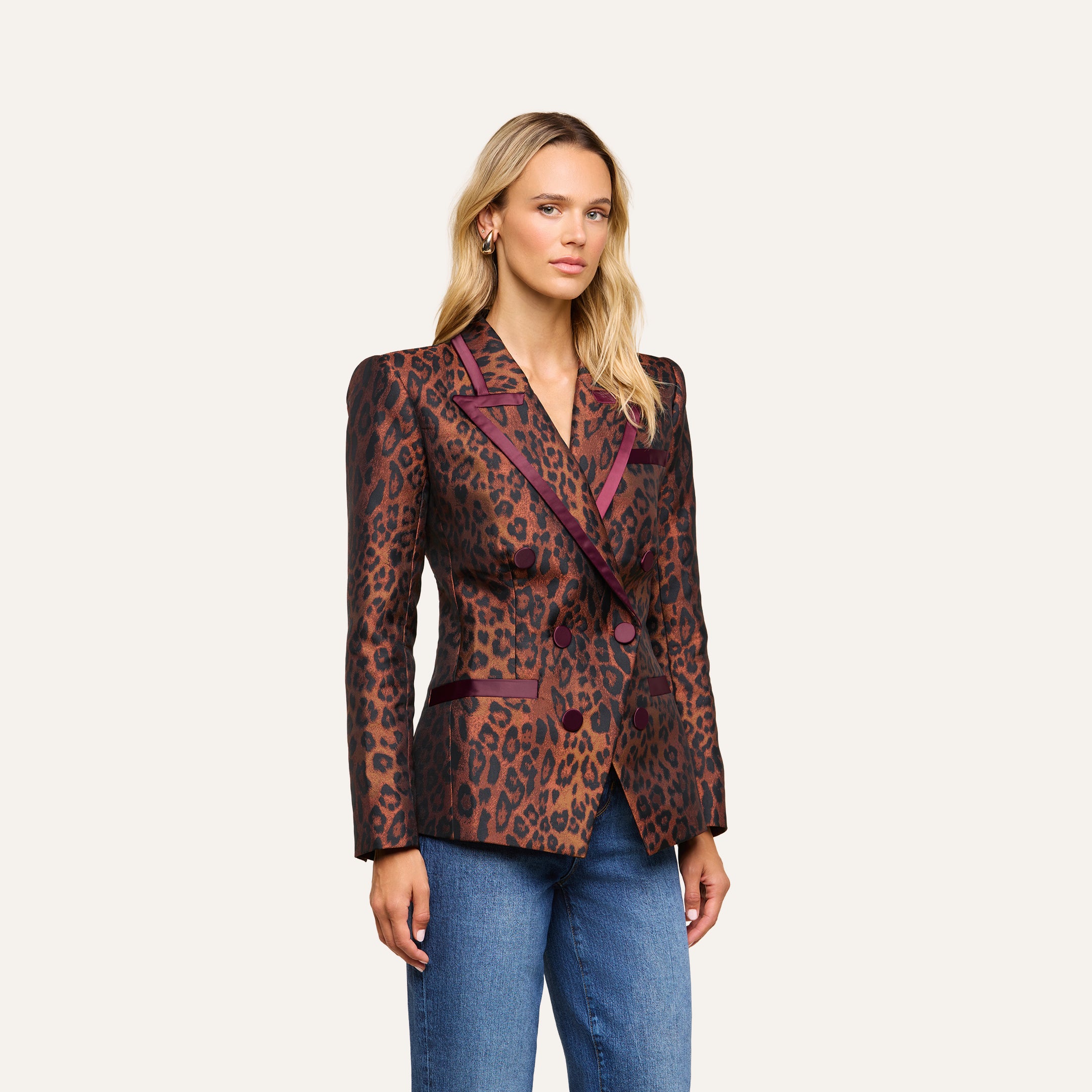 A woman with long blonde hair wears the Kyomi Double-Breasted Blazer—a fitted, longline leopard jacquard jacket with dark accents—paired with blue jeans, standing against a plain light background.