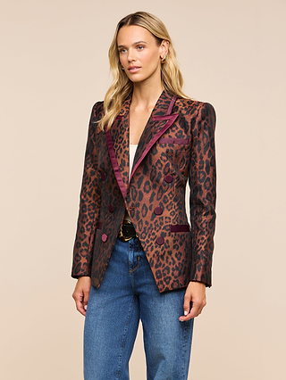 A woman with wavy blonde hair wears the Kyomi Double-Breasted Blazer, featuring a leopard jacquard pattern with burgundy accents, over a white top and blue jeans, as she stands against a beige background.