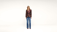 A person wearing sunglasses, a patterned blazer, blue jeans, and heels stands confidently against a plain white background.