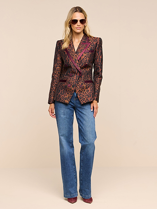 A woman wearing sunglasses, the Kyomi Double-Breasted Blazer in a leopard print with maroon details, blue jeans, and maroon shoes stands against a plain beige background, facing forward with a relaxed posture.