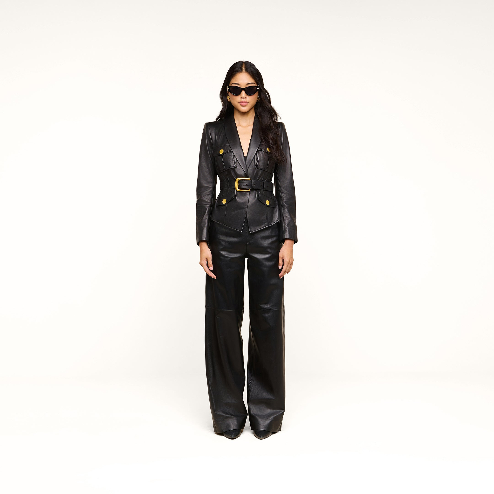 A woman wearing the Wisteria Leather Blazer with gold buttons, wide-leg black leather pants, and sunglasses stands against a plain white background.