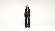 A person wearing black sunglasses, a black belted blazer, and black wide-leg pants stands against a plain white background.